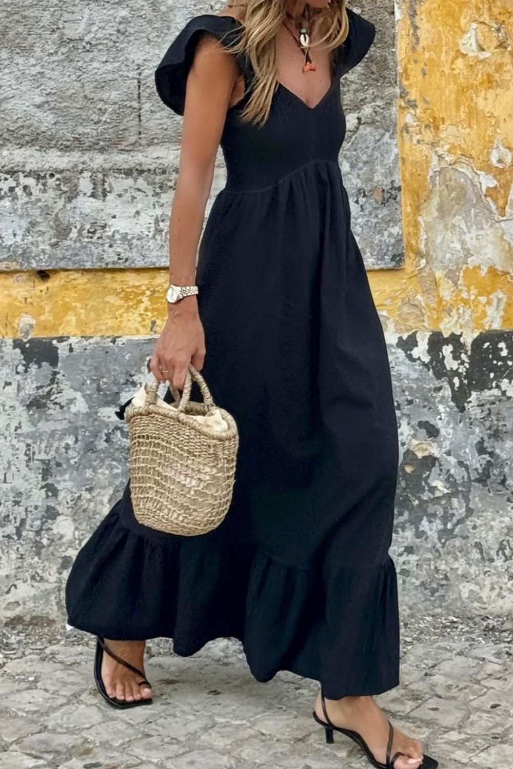A woman wearing a black maxi dress with ruffled cap sleeves and a smocked waist, paired with black heels and carrying a straw tote bag.