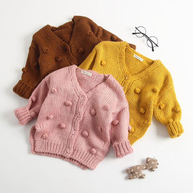 Three baby sweater cardigans in pink, brown, and yellow colors, displayed on a white background, with a pair of glasses and a small toy car visible in the corner.