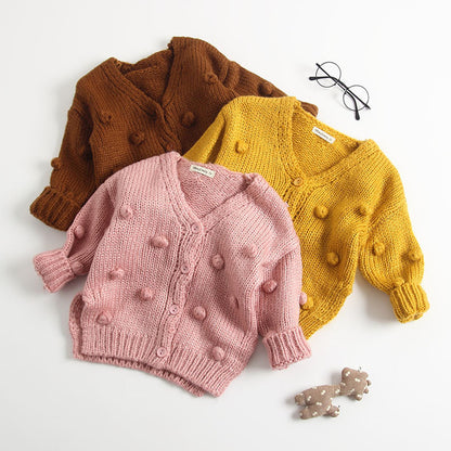 Three baby sweater cardigans in pink, brown, and yellow colors, displayed on a white background, with a pair of glasses and a small toy car visible in the corner.